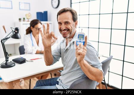 Ein Mann mittleren Alters in der Arztpraxis hält das Blutzuckermessgerät in der Hand und macht ein gutes Zeichen mit den Fingern, lächelnd freundlich Gesten ausgezeichnetes Symbol Stockfoto
