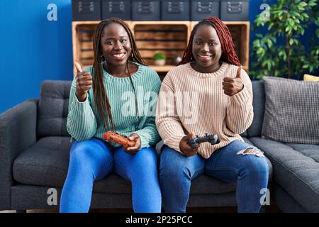 Zwei afrikanische Frau, die Videospiele spielt, mit einem Controller, der glücklich und positiv lächelt, Daumen hoch, ausgezeichnet, und Bestätigung unterschrieben Stockfoto