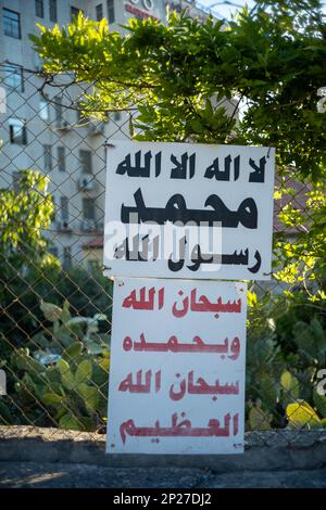 Das weiße Schild auf Arabisch auf einem Zaun bei Einem Park steht: "Es gibt keinen Gott außer Allah. Muhammad ist der Bote Gottes. Ruhm sei Allah, und möge er gelobt werden. Stockfoto