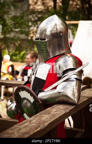 Kostüm aus der Mittelalterzeit beim Ritterturnier. Mittelalterliche historische Nachstellung - ein Mann mit Metallhelm und roter Rüstung, mit einem Schwert und Stockfoto