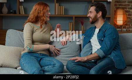 Frau, die zu Hause mit einem Mann spricht fröhliches weißes Paar sitzt auf der Couch rothaariges Mädchen erzählt lustige Geschichte positive, vielfältige Partner lachen mit Witz Stockfoto