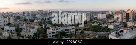 Breites Stadtbild von Ramallah bei Sonnenaufgang mit Sonnenuntergang, hohen Gebäuden und Bäumen vor blauem und violettem Himmel Stockfoto