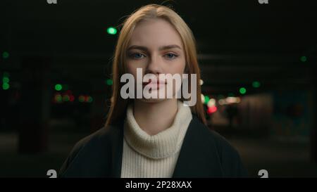 Eine ruhige Weiße steht allein auf dem dunklen Parkplatz. Fokussiertes blondes Mädchen, das auf die Kamera schaut. Porträt einer Geschäftsfrau Stockfoto