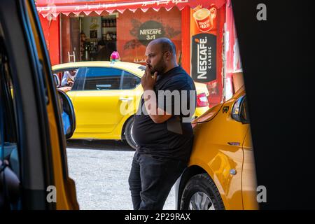 Bethlehem, Westjordanland, Palästina - 22. Juli 2022: Der große Glatzkopf-Araber im schwarzen Hemd raucht eine Zigarette. Er lehnt sich auf Ein gelbes Taxi, das vor einem Nesc geparkt ist Stockfoto