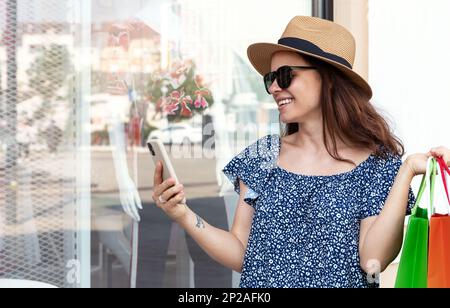 Eine Frau mit Strohhut, die Einkaufstaschen in der Hand hält und auf ihr Smartphone schaut. Stockfoto