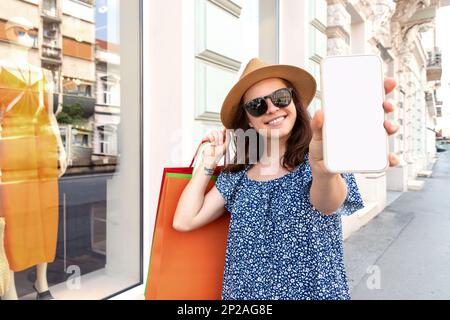 Frau mit Einkaufstasche, die einen leeren Bildschirm ihres Handys in die Kamera zeigt und lächelt. Stockfoto