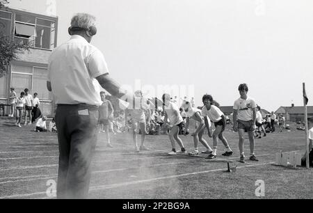 1989, Schulsporttag, draußen auf einem Sportplatz, männliche Schüler der Sekundarstufe bereit für ein Rennen, als männlicher Lehrer mit Gehörschutz, der Starter, wenn Rauch erscheint, feuert die Startpistole, England, Großbritannien. Stockfoto