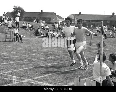 1989, ein Sporttag an einer Sekundarschule, zwei männliche Jugendliche, die an einem Laufrennen auf der Rasenbahn eines Sportplatzes teilnehmen, Sleaford, Lincs, England, Großbritannien. Ein Schuljunge, der auf dem Rasen sitzt, hält eine Rundennummer. Stockfoto