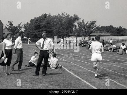 1989, Schulsporttag an einer Sekundarschule, ein männlicher Teenager, der an einem Rennen auf der Rasenbahn teilnimmt, mit einer Lehrerin, die nebenan steht und die Runden zählt, Sleaford, England, Großbritannien. Stockfoto