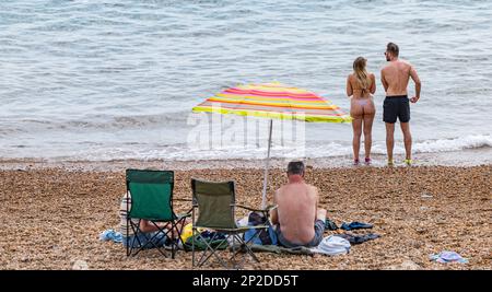 Ein Paar am Strand mit einer jungen Frau, die einen Bikini-Badeanzug am Seatown Beach in Summer an der Jurassic Coast, Dorset, England, UK trägt Stockfoto