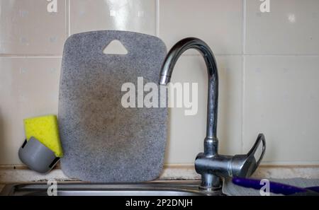 Wasserhahn und graues Schneidebrett im Hintergrund. Dreckiges Schneidebrett in der Nähe des Waschbeckens, ungewaschenes Geschirr nach dem Kochen des Abendessens. Tägliche Aufgaben. Stockfoto