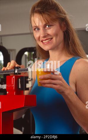 Eine junge Frau in einem Fitnessstudio, die nach dem Training pausiert, um auf die Waage zu steigen und sich selbst zu wiegen, während sie ein Glas Orangensaft in der Hand hält Stockfoto