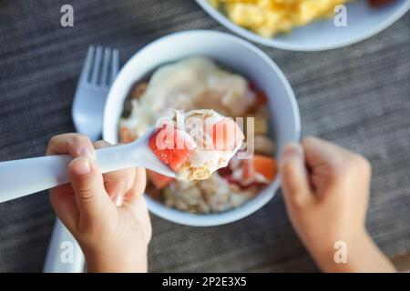 Ein kleines Mädchen isst Müsli mit Joghurt und Obst mit weißem Plastiklöffel Stockfoto