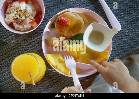 Kindermenü - Omelett, Pfannkuchen mit Ahornsirup, Müsli mit Joghurt und ein Glas frisch gepressten Orangensaft auf dem Restauranttisch mit Plastik Stockfoto