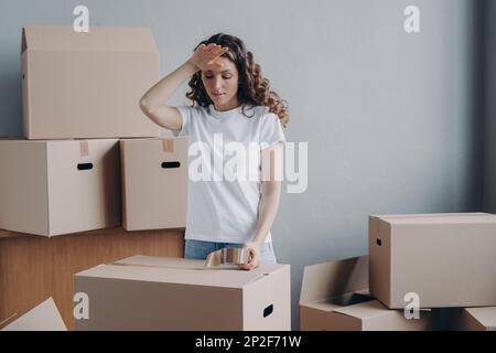 Müde Frau, die Kartons mit Sachen packt, Klebeband benutzt, um Schweiß von ihrer Stirn zu wischen. Überarbeitete, erschöpfte spanierin, die sich darauf vorbereitet Stockfoto