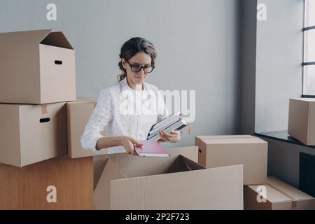 Eine Frau trägt Brille, die am Umzugstag ein Notebook mit Pappkartons packt, eine fokussierte hispanische Geschäftsfrau packt Dinge in Karton, Prepar Stockfoto