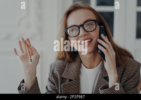 Lächelnde weibliche Berufsanruferin, die im Büro telefoniert und einen Bleistift in der Hand hält. Eine Brille tragende Geschäftsfrau beantwortet einen Anruf und kommuniziert mit ihr Stockfoto