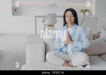 Ruhige Frau übt Yoga, meditiert, sitzt in Lotus-Pose zu Hause auf dem Sofa. Die Frau sitzt auf der Couch und entspannt die gefalteten Hände in einer namensfreundlichen Geste. Gut Stockfoto