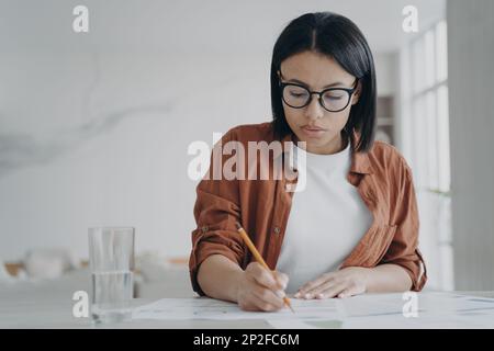 Fokussierte Geschäftsfrau mit Brille arbeitet an einem geschäftlichen Projekt, macht Notizen, sitzt am Arbeitsplatz. Moderne weibliche Managerin, die arbeitet, schreibt Stockfoto