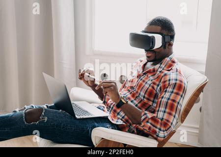 Ein moderner afroamerikanischer Mann mit einer VR-Brille, der im Cyberspace auf einem Laptop arbeitet, ein schwarzer Entwickler oder Designer, der mit der virtuellen Realität interagiert Stockfoto