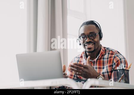 Ein afroamerikanischer Mann, der ein Headset trägt, kommuniziert mit dem Kunden per Videoanruf über einen Laptop, der im Home Office am Schreibtisch sitzt. Freundlicher junger schwarzer Hotlin Stockfoto