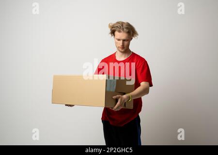 Der Zusteller in roter Uniform trägt einen Pappkarton in den Händen auf weißem Hintergrund. Der Zusteller übergibt die Paketlieferung. Stockfoto