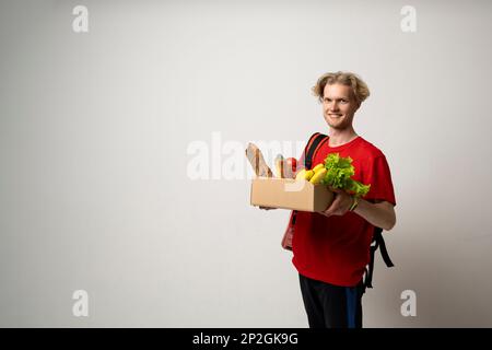Happy Delivery Mann in roter Uniform mit einer Kiste voller Lebensmittel auf weißem Hintergrund. Express-Lieferung, Food-Lieferung, Online-Shopping-Konzept. Stockfoto