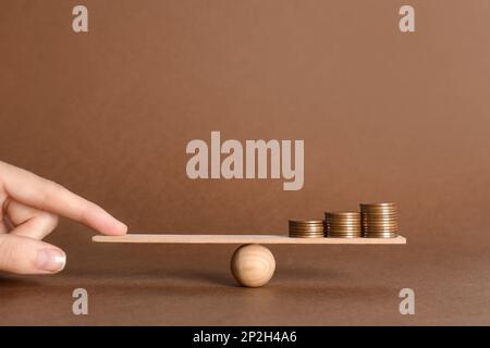 Frau balanciert Miniatur-Wippe mit Münzstapeln auf braunem Hintergrund, Nahaufnahme. Harmoniekonzept Stockfoto