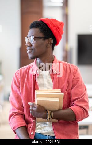 Junger, glücklicher afroamerikanischer Universitätslehrer, der im Klassenzimmer sitzt und einen Stapel Bücher hält Stockfoto