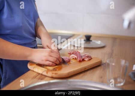Fleisch auf einem Brett schneiden, Frauenhände mit einem Messer. Unverstellter natürlicher Schuss einer Frau beim Kochen in der Küche Stockfoto