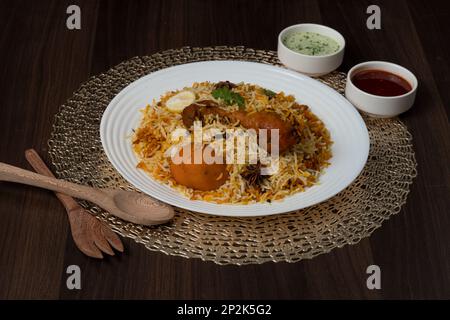 Huhn Biryani mit Saucen, beliebte pakistanische indische Gerichte auf einem weißen Teller auf dunklem strukturiertem Hintergrund. Stockfoto
