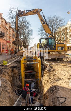 Grodno, Weißrussland - 06. März 2022: Hydraulikbagger füllt Tiefaushub und wird während der Installation des Abflussrohrs von einem Grabenkasten mit Erbsenschieferkies unterstützt Stockfoto