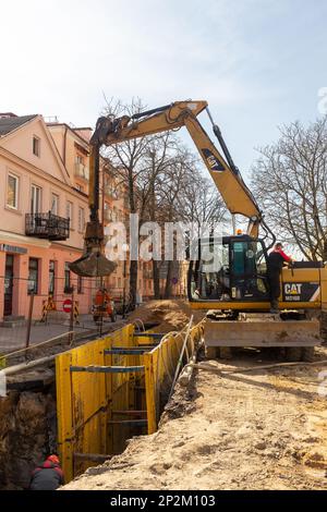 Grodno, Weißrussland - 06. März 2022: Hydraulikbagger füllt Tiefaushub und wird während der Installation des Abflussrohrs von einem Grabenkasten mit Erbsenschieferkies unterstützt Stockfoto