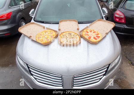 Drei Pizzas in offenen Pappkartons liegen auf der Motorhaube eines Autos auf dem Parkplatz. Ein Set aus drei Pizzen zum Mittagessen für eine Gruppe von Reisenden Stockfoto