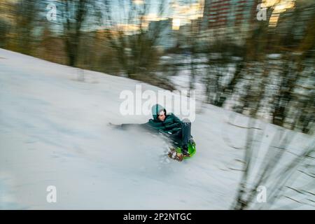 02-12-2022 Moskau Russland. Ein junger Mann, der auf einem Plastikschild von den Krylatsky-Hügeln in Moskau herunterrollt. Winter Februar in Russland. Stockfoto