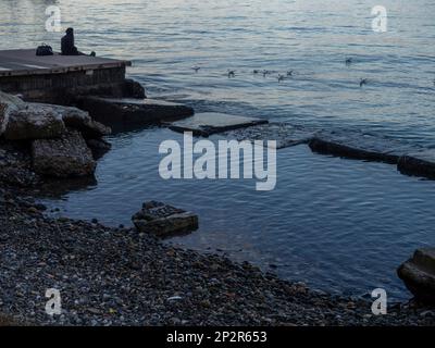 Ein einsames Mädchen sitzt auf einem Pier im Hafen. Er sitzt am Rand des Wassers. Konzept der Einsamkeit. Stockfoto