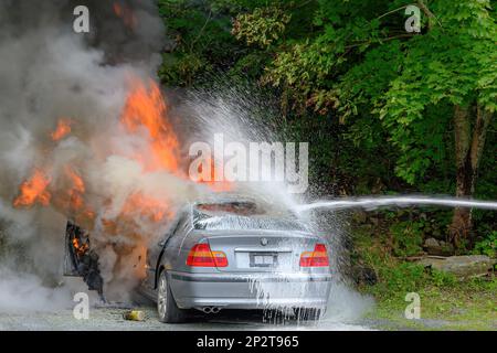 Saint John, NB, Kanada - 18. August 2022: Ein Auto, das in Flammen stand. Ein Wasserstrahl aus Schaum bekämpft das Feuer von rechts. Bäume im Hintergrund. Ge Stockfoto