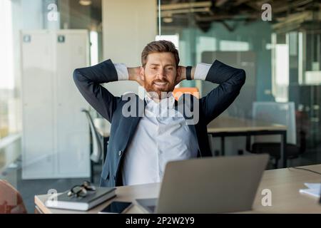 Glücklicher, entspannter Geschäftsmann, der eine Pause von der Arbeit macht, auf einem Stuhl sitzt und Hände hinter dem Kopf hält Stockfoto