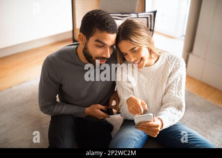 Die freundliche Millennials-europafrau zeigt dem araber ihr Smartphone und liest Nachrichten auf dem Boden im Zimmer Stockfoto