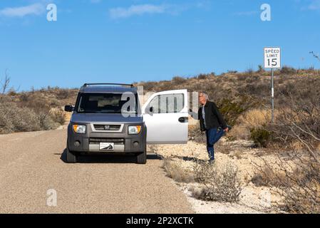 Auf einer Autoreise hält ein 80-jähriger Mann an, um sich ein Bein zu strecken, während er sich an der Tür seines Honda Elements festhält. Stockfoto