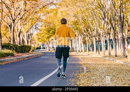 Ein Mann läuft, joggt draußen auf der Straße mit Herbstbäumen mit gelben Blättern auf beiden Seiten der Straße Stockfoto