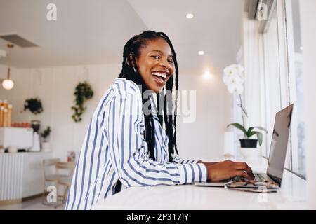 Junge, ehrgeizige Freiberuflerin, die glücklich lacht, während sie in einem Café an einem Laptop arbeitet. Fröhliche junge schwarze Geschäftsfrau, die online in einem Kaffee arbeitet Stockfoto