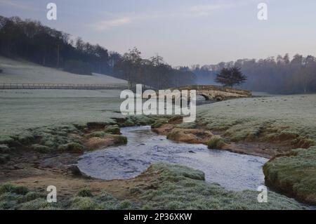 Blick auf die gewölbte Brücke über einen Bach, der im Morgengrauen durch die Parklandschaft fließt, Hovingham Park, Hovingham, North Yorkshire, England, Vereinigtes Königreich Stockfoto