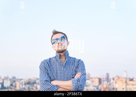 Weißer Mann in Schlafkleidung und Superhelden blaue Maske steht draußen mit ernstem Gesicht. Urbane Ansicht im Hintergrund. Hochwertige Bilder Stockfoto
