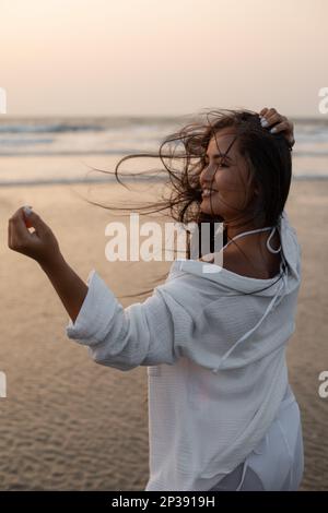 Junge, schöne, lächelnde Brünette mit lockeren langen Haaren steht umgedreht am Meer vor Sonnenuntergang. Stockfoto