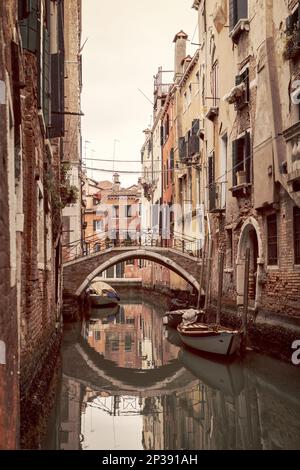 Gewölbte Brücke über einen engen Kanal in Venedig mit alten Häusern und geparkten Booten daneben (Foto im Vintage-Stil) Stockfoto