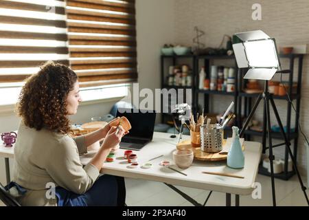 Töpferinnen in blauer Schürze machen einen Vlog, nehmen Online-Kurs auf, Lehm-Master-Klasse, Unterricht in ihrem Studio mit Steingut-Regal im Hintergrund Stockfoto