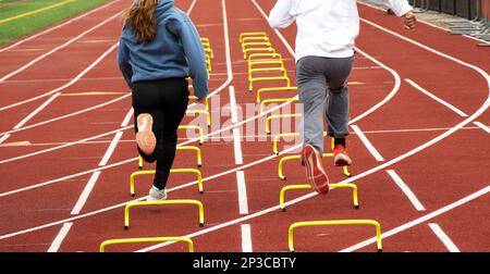 Rückansicht von zwei Leichtathleten, die beim Training auf einer kalten Rennstrecke über kleine gelbe Hürden in der Spur laufen. Stockfoto