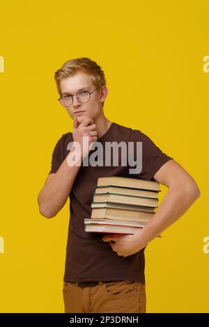 Wissenserwerb, Ausbildung, Konzeptschulung. Ein grüblerischer junger Europäer mit braunem T-Shirt hält einen Stapel Bücher auf gelbem Hintergrund in der Hand. Hochwertiges Foto Stockfoto