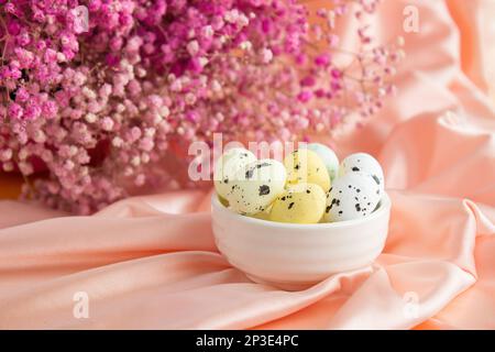 Osterzusammensetzung auf dem Hintergrund rosa Satinwachteleier in einer weißen Keramikschüssel. Auf der Rückseite ist ein Strauß rosa Zigeuner Stockfoto